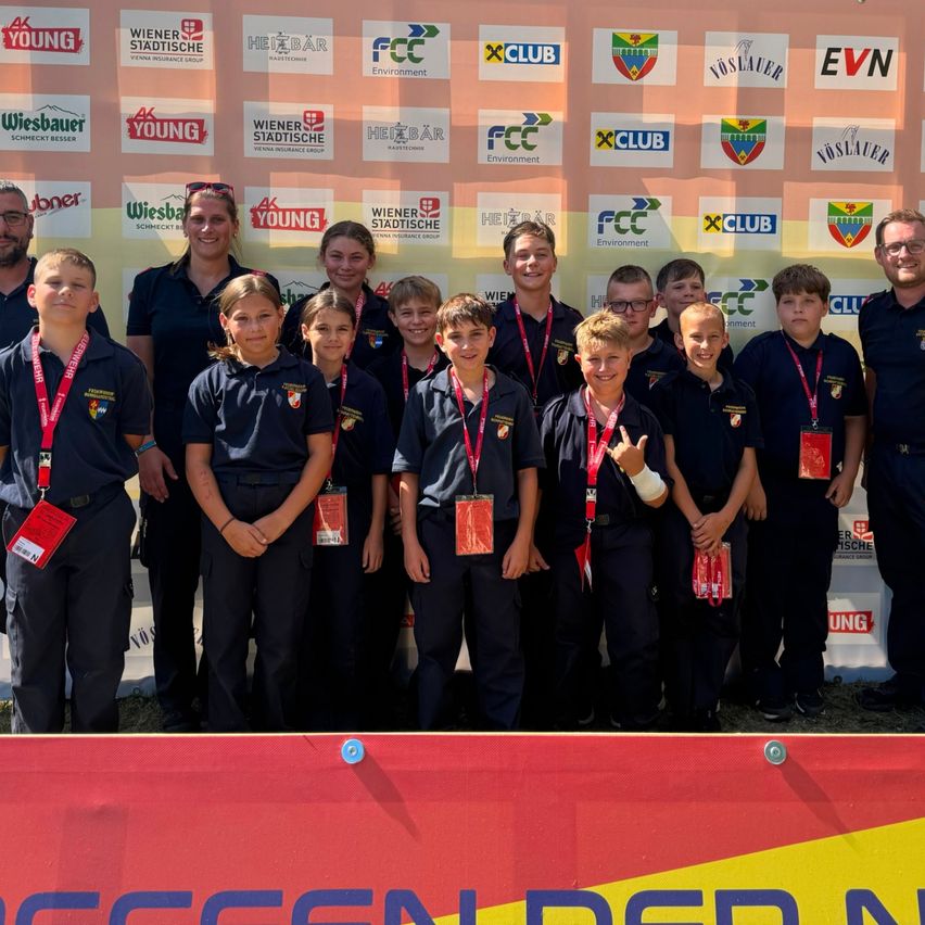 Eine Gruppe von Kindern und Erwachsenen in Marineblau-Uniformen mit roten Namensschildern posiert für ein Foto vor einer Wand mit verschiedenen Logos.