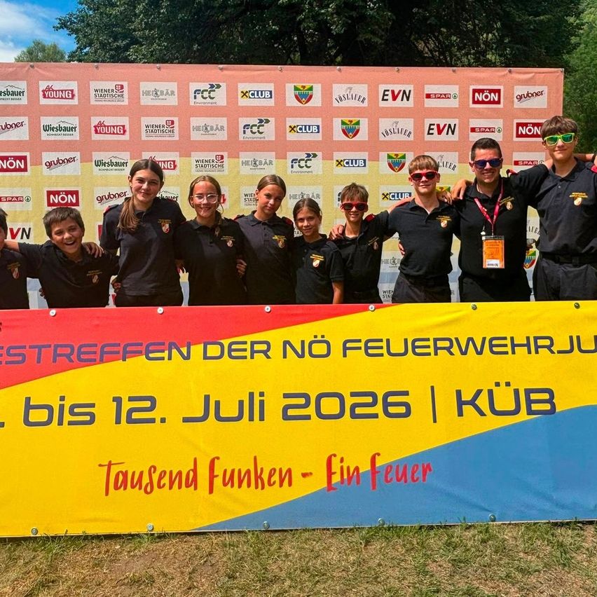 Eine Gruppe junger Menschen in Uniform steht vor einem Banner, das eine Jugendfeuerwehrveranstaltung im Juli 2026 bewirbt. Sie lächeln und posieren für ein Foto.