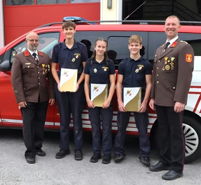 Fünf Personen stehen vor einem roten und weißen Van, möglicherweise einem Feuerwehrwagen. Sie sind alle in Uniform gekleidet und halten jeweils ein Zertifikat.