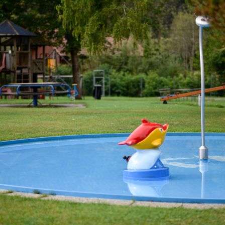 Ein kleiner Pool mit einer roten und gelben Fischfigur auf einer blauen Basis befindet sich in einem grünen Park mit einem Spielhaus und Rutschen.