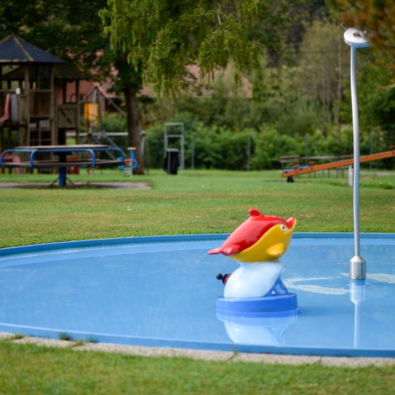 Ein kleiner Pool mit einer roten und gelben Fischfigur auf einer blauen Basis befindet sich in einem grünen Park mit einem Spielhaus und Rutschen.
