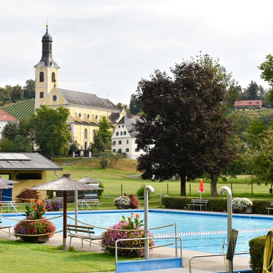 Ein Freibad ist von Bänken und Blumen umgeben. Eine große Kirche mit einem Glockenturm ist in der Ferne. Bäume und Büsche sind in der Nähe. Der Himmel ist bewölkt.