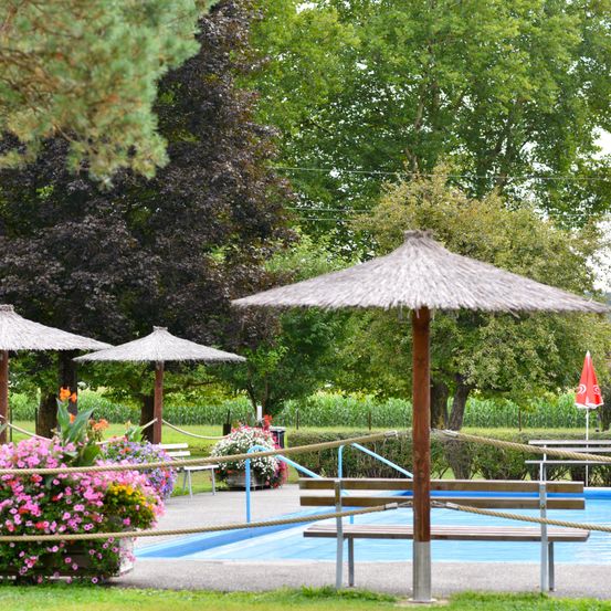 Ein von Grün und Blumen umgebener Pool. Es gibt mehrere strohgedeckte Sonnenschirme, einen geschlossenen roten Schirm und Bänke. Bäume und Büsche sind im Hintergrund.