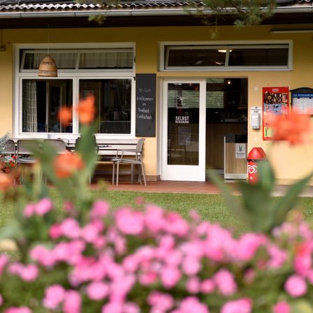 Ein gelbes Gebäude mit einem Schild, das 'Treadmill Lauf' liest. Ein Schreibtisch, Stühle und Blumen sind draußen. Die Tür hat eine Glasscheibe. Das Gebiet hat einen Rasen.
