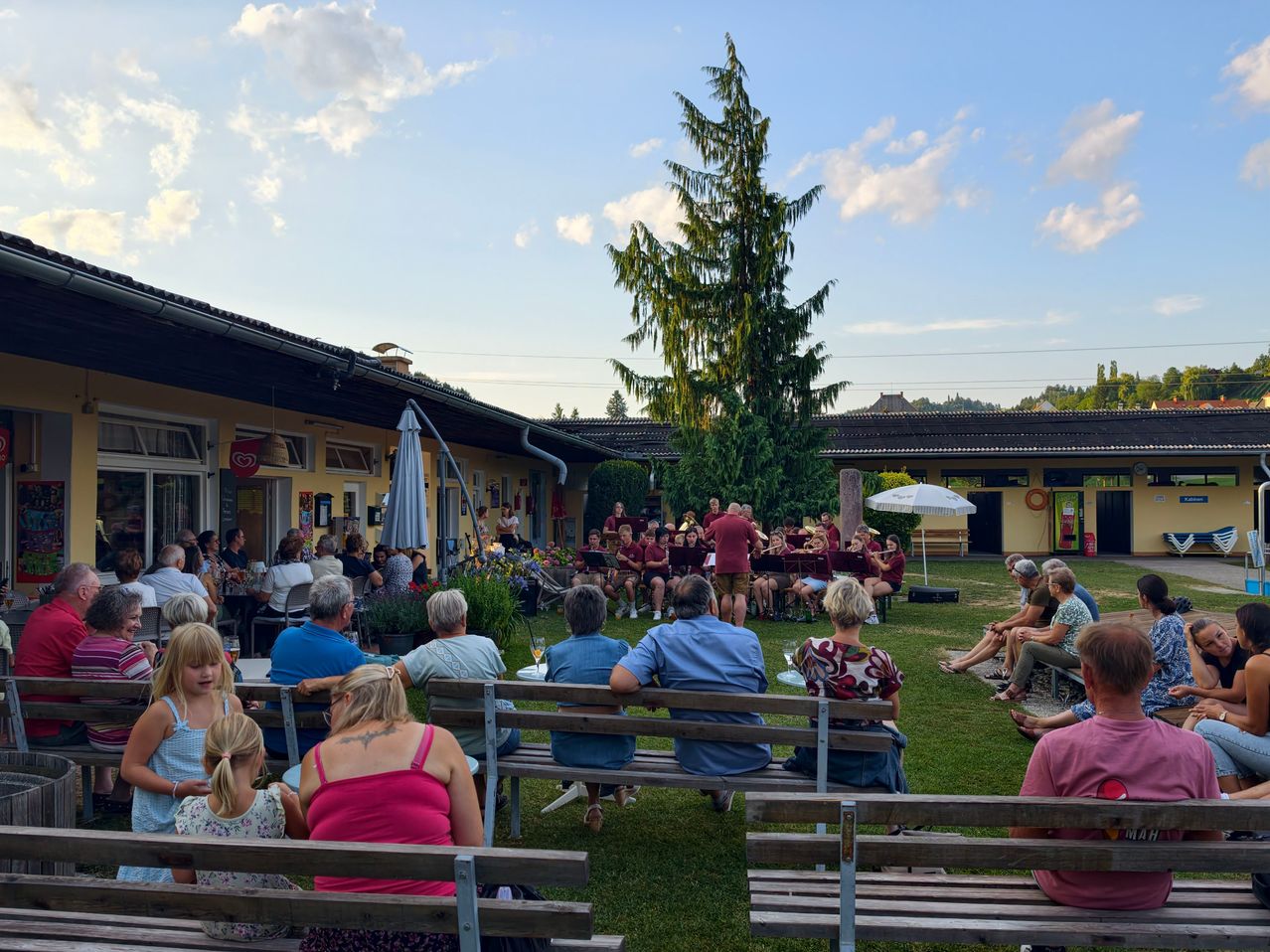 A group of people are sitting on benches in a park, watching a band play on a stage. The band is dressed in red uniforms.