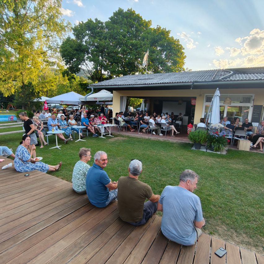 A group of people are sitting on a wooden deck in front of a building with a grassy lawn. Some are seated while others are standing. There are tables and chairs set up, and a pool is visible in the background.