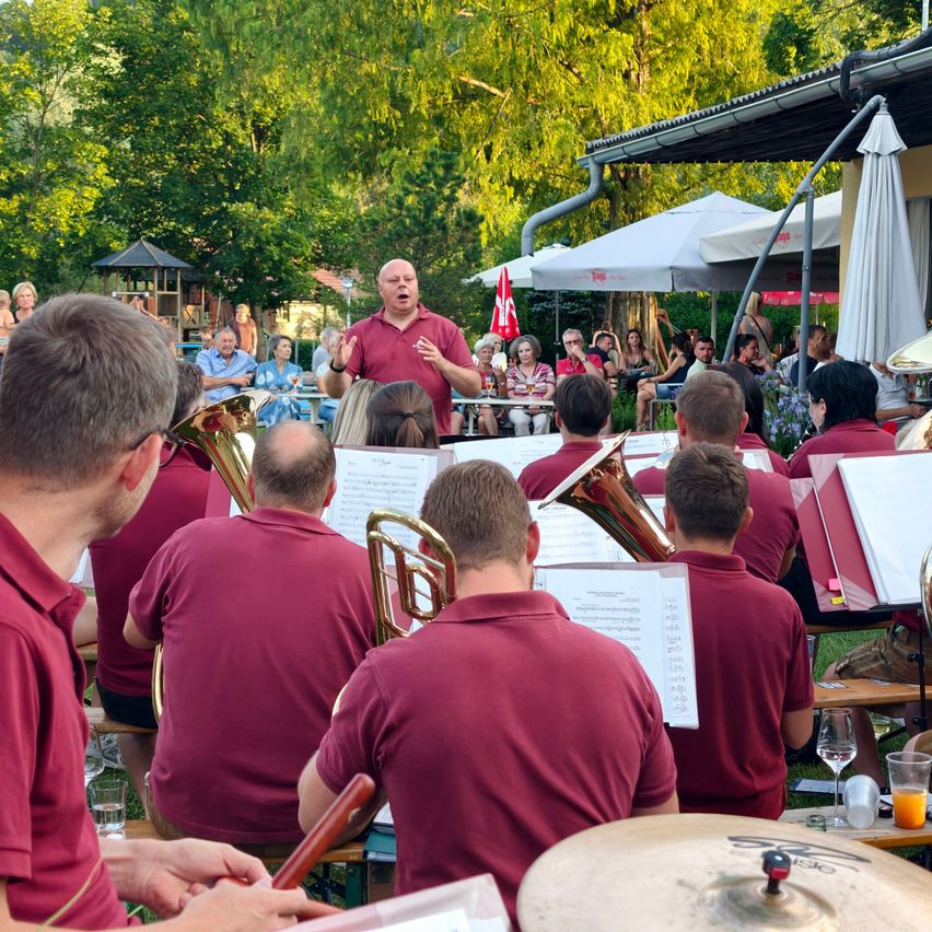 A band performs on an outdoor stage with a conductor leading, while an audience watches from the surrounding area.