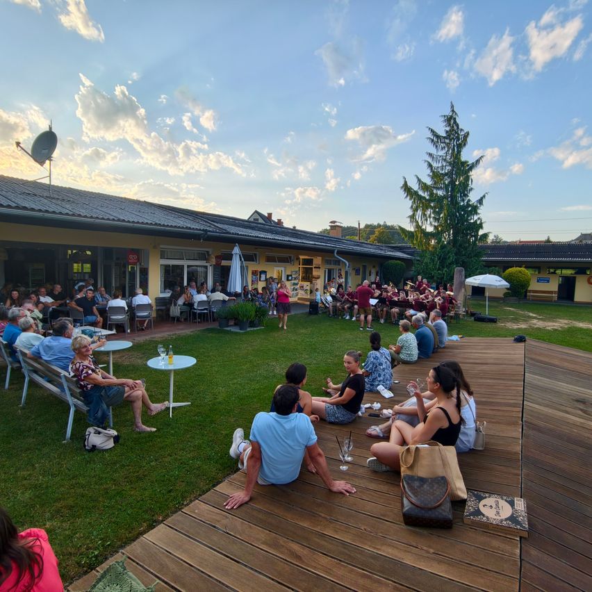 A group of people are seated on a wooden deck in front of a building, watching a band perform on a grassy lawn.