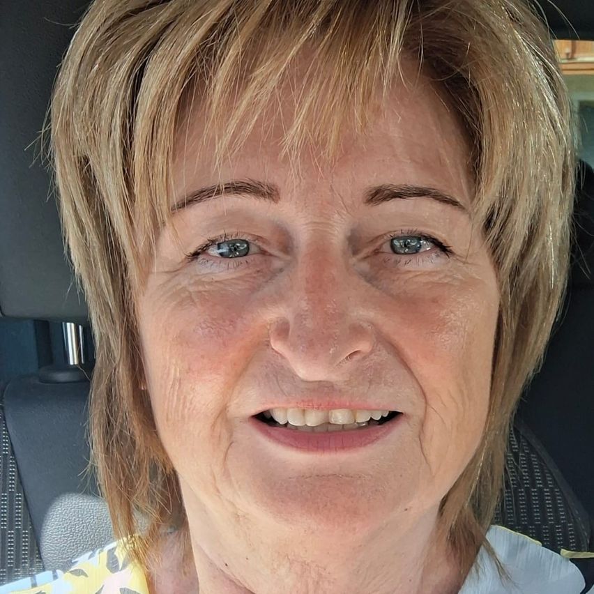 Eine ältere Frau mit kurzen blonden Haaren und blauen Augen lächelt. Sie sitzt in einem Auto und trägt ein Blumenhemd.