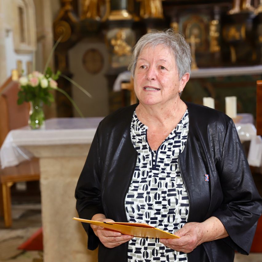Eine ältere Frau steht in einer Kirche und hält ein Buch. Hinter ihr befindet sich ein Altar mit Blumen und Kerzen.