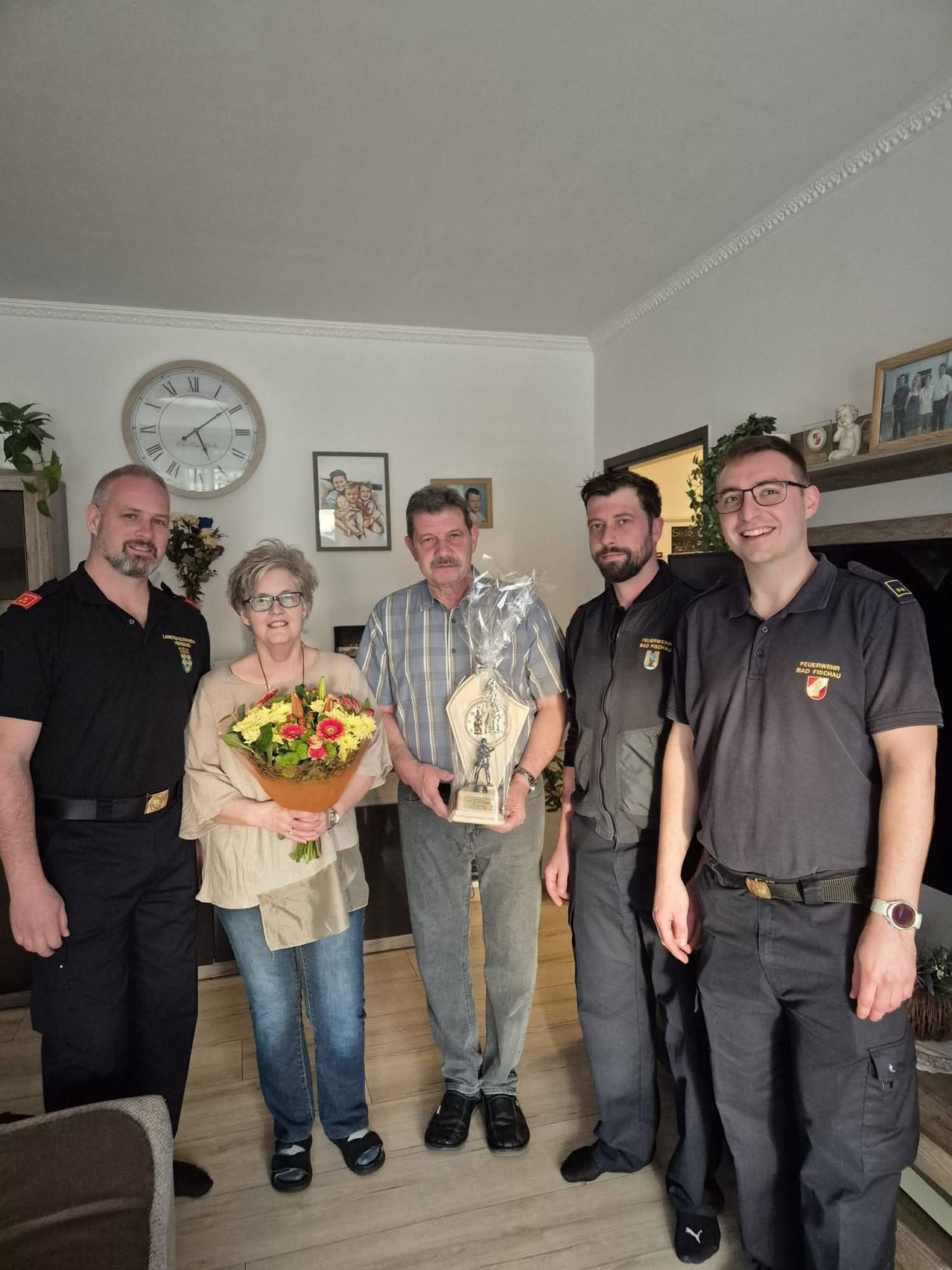 Fünf Personen stehen lächelnd zusammen im Innenraum. Der Mann in der Mitte hält eine Trophäe, während die Frau einen Blumenstrauß hält. Dahinter sind eine Uhr, gerahmte Bilder und ein Regal zu sehen.