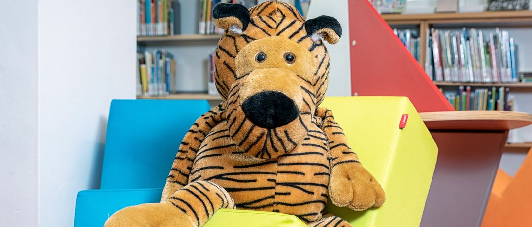 Ein großer, getigerter Plüschtier sitzt auf einem gelben Sechseckkissen in einer Bibliothek mit bunten Stühlen und mit Büchern gefüllten Bücherregalen.