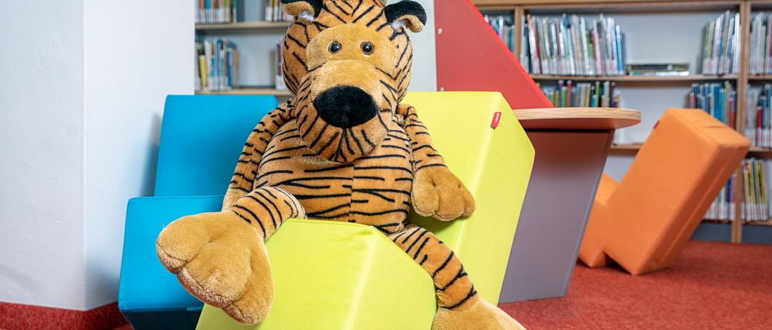 Ein großer, gestrickter Tiger sitzt auf einem gelben Würfel in einem farbenfrohen Raum, mit Bücherregalen und einem Schreibtisch im Hintergrund.
