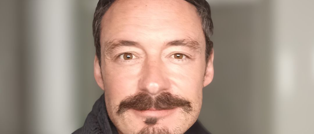 Ein Mann mit Bart und Schnurrbart lächelt und trägt ein gelbes Shirt und eine schwarze Jacke.