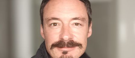 Ein Mann mit Bart und Schnurrbart lächelt und trägt ein gelbes Shirt und eine schwarze Jacke.