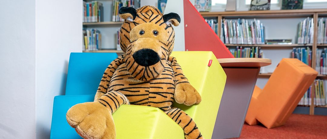 Ein Tigerplüsch sitzt auf einem gelben Block in einem hell beleuchteten Raum mit farbenfrohen Möbeln und mit Büchern gefüllten Bücherregalen.