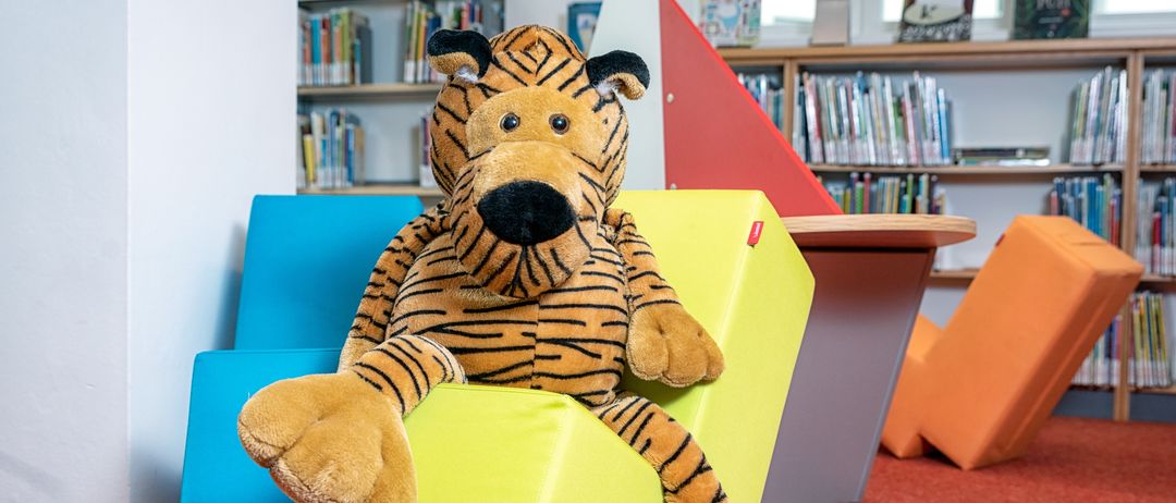 Ein gedönstiger Tiger sitzt auf einem leuchtend gelben Würfel in einer Bibliothek mit Regalen voller Bücher.