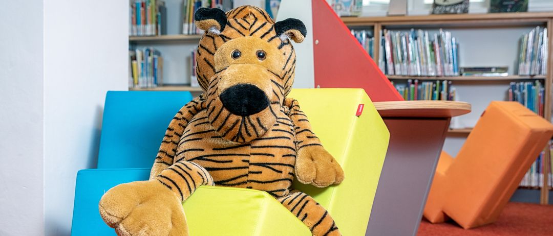 Ein großer pelziger Tiger sitzt auf einem gelben Sechseck-Kissen in einer Bibliothek, umgeben von bunten Bausteinen und Bücherregalen voller Bücher.