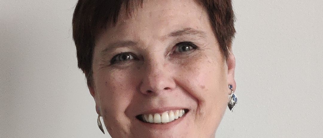 Eine lächelnde Frau mit kurzen Haaren, die eine blaue Jacke und eine Halskette mit einem blauen Stein trägt. Sie posiert für ein Foto.