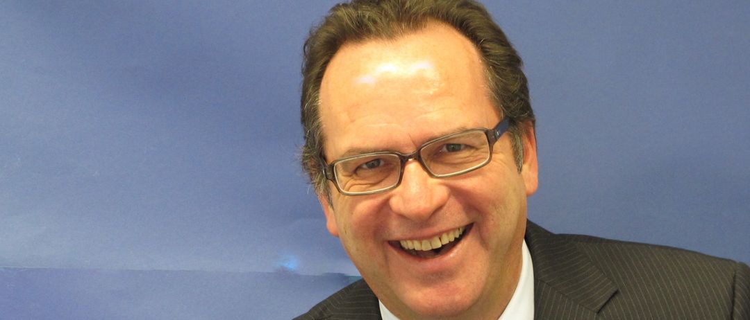 Ein Mann in Anzug und Krawatte lächelt für ein Foto vor blauem Hintergrund.