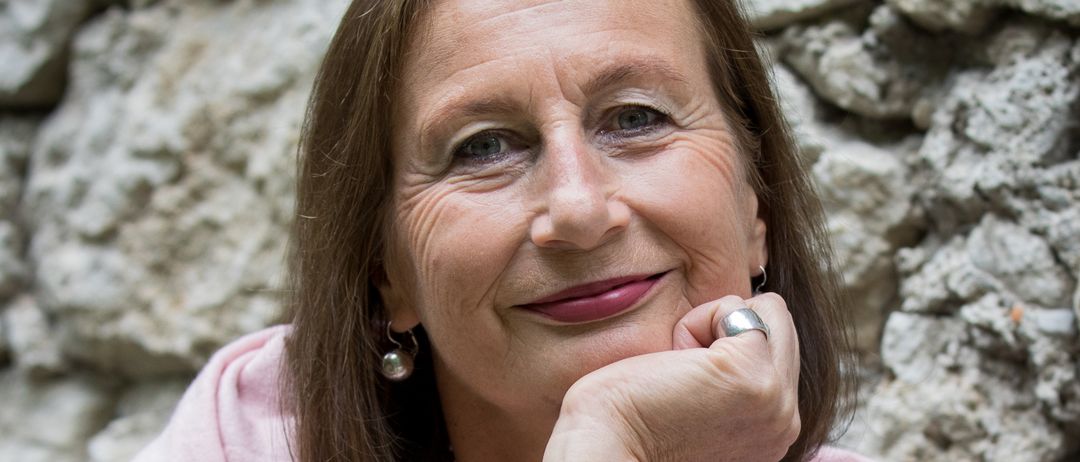 Eine ältere Frau mit braunen Haaren, die einen rosa Schal und ein graues Hemd trägt, lächelt und posiert vor einer Steinmauer. Sie hat einen silbernen Ring und eine Uhr an ihrer linken Hand.