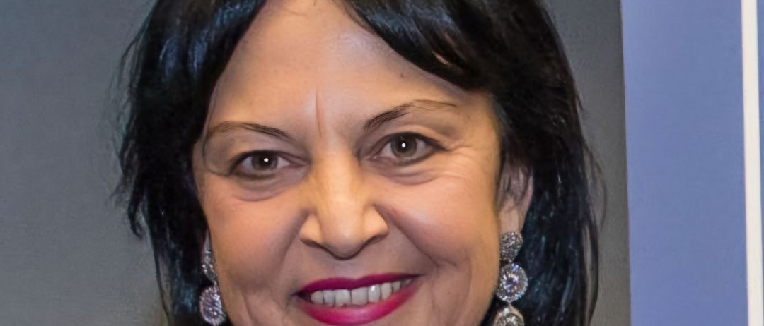 Eine Frau mit schwarzen Haaren lächelt und posiert für ein Foto. Sie trägt ein lilafarbenes Oberteil, eine Halskette und große Ohrringe. Sie befindet sich wahrscheinlich in einer Indoor-Umgebung mit einem blauen Hintergrund.