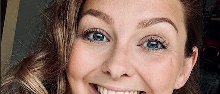 Eine Frau mit langen, gewellten braunen und blonden Haaren lächelt in die Kamera und trägt ein weißes T-Shirt. Sie hat blaue Augen und einen Muttermal auf ihrer Wange.