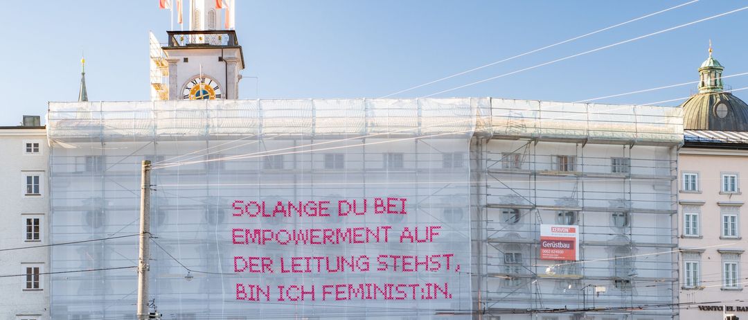 Ein großes Gebäude im Bau mit Gerüst und Kabeln. Ein Uhrturm steht darüber mit Flaggen. Ein deutscher Slogan ist auf der Gebäudeseite gemalt.