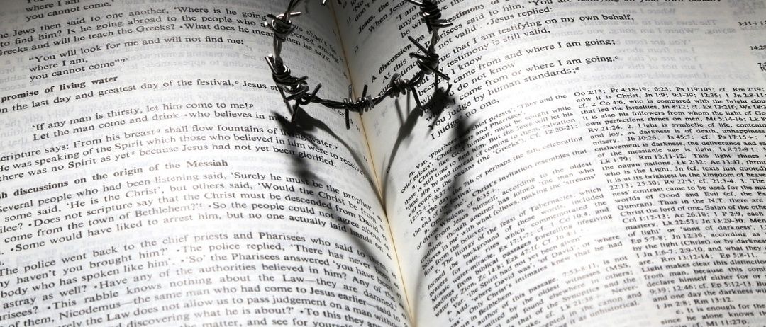Ein offenes Buch mit einer Dornenkrone darauf, die einen Schatten wirft. Die Seite zeigt einen religiösen Text, möglicherweise aus der Bibel.