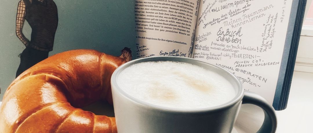 Ein offenes Buch mit dem Titel 'Der Mann, der noch an den Klapperstorch glaubte' mit einem Croissant und einer Tasse Kaffee in der Nähe.