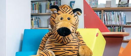 Ein großer gedönster Tiger sitzt auf einem hellgelben Würfel in einem farbenfrohen Raum mit Bücherregalen im Hintergrund.