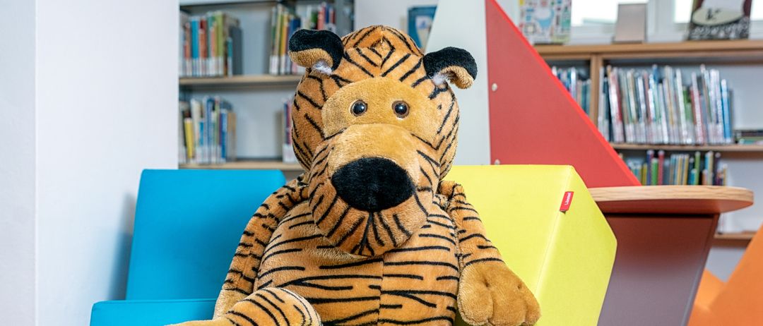Ein großer gedönster Tiger sitzt auf einem hellgelben Würfel in einem farbenfrohen Raum mit Bücherregalen im Hintergrund.