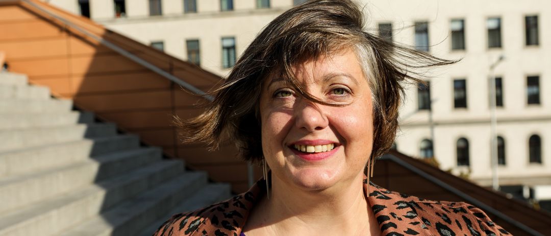 Eine Frau mit kurzen Haaren lächelt vor einem Gebäude mit einer Treppe und moderner Architektur. Sie trägt eine Leoparden-Jacke über einem lila Shirt.