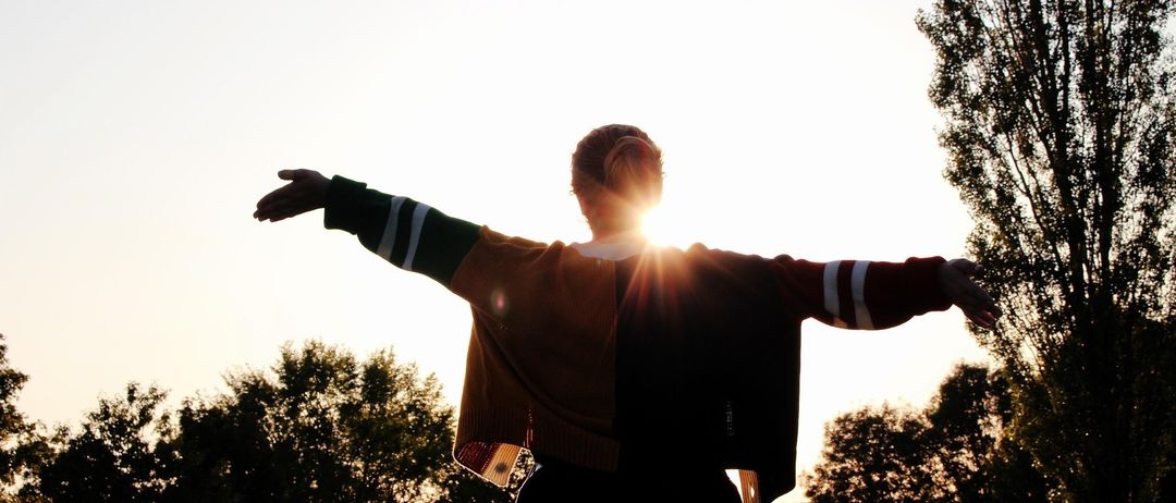 Eine Person steht mit ausgestreckten Armen in einem Feld, von der untergehenden Sonne beschienen.