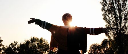 Eine Person steht mit ausgestreckten Armen in einem Feld, von der untergehenden Sonne beschienen.