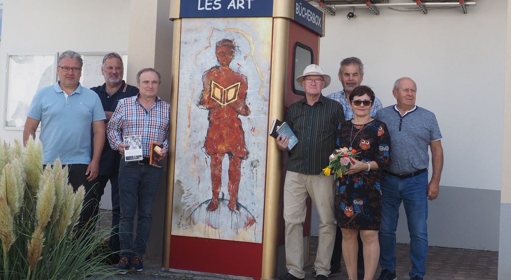 Eine Gruppe von Menschen posiert für ein Foto vor einer bemalten Telefonzelle mit einem Vogel obenauf und den Worten Les Art Bucherbox.