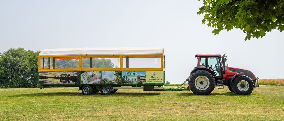 Ein roter Traktor zieht einen gelben Anhänger mit weißem Dach auf einem Feld unter klarem Himmel. Es gibt Bilder an der Seite des Anhängers.
