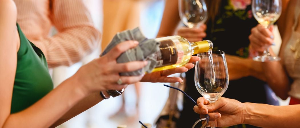 Eine Gruppe von Menschen versammelt sich, wobei eine Person Weißwein in ein Glas gießt, das von einer anderen Person gehalten wird. Im Hintergrund sind weitere Personen zu sehen, die Weingläser halten.