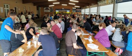 Viele Menschen sitzen an Tischen in einem großen Raum. Sie unterhalten sich und einige tragen Brillen. Der Raum hat Holzdecken und Wände, und Poster sind an den Wänden angebracht. Es gibt Tische mit Flaschen, Gläsern und anderen Gegenständen darauf. Der Raum hat große Fenster auf einer Seite.
