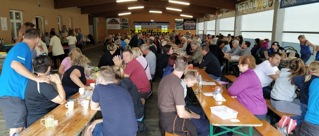 Viele Menschen sitzen an Tischen in einem großen Raum. Sie unterhalten sich und einige tragen Brillen. Der Raum hat Holzdecken und Wände, und Poster sind an den Wänden angebracht. Es gibt Tische mit Flaschen, Gläsern und anderen Gegenständen darauf. Der Raum hat große Fenster auf einer Seite.