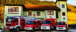 Mehrere Feuerwehrfahrzeuge sind vor einem Gebäude mit dem Namen Dorf geparkt. Die Fahrzeuge sind rot und weiß, eines davon hat eine Leiter oben auf. Im Hintergrund ist ein klarer Himmel mit hohen Bäumen.