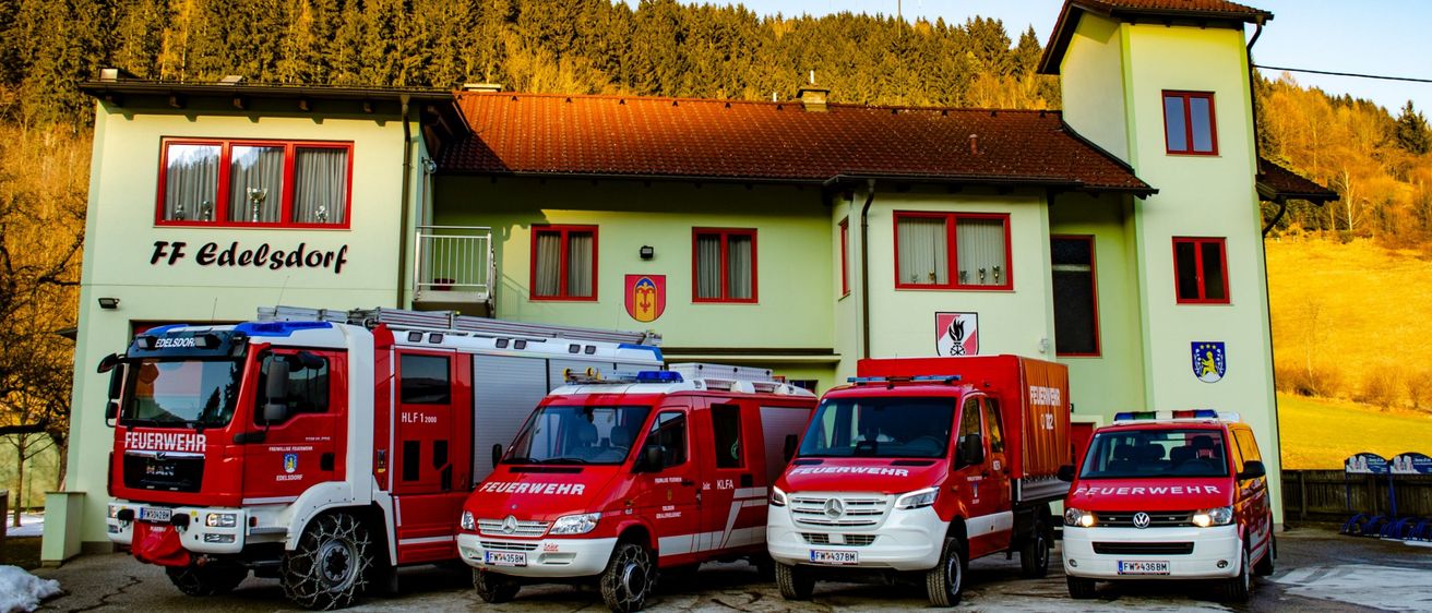 Mehrere Feuerwehrfahrzeuge sind vor einem Gebäude mit dem Namen Dorf geparkt. Die Fahrzeuge sind rot und weiß, eines davon hat eine Leiter oben auf. Im Hintergrund ist ein klarer Himmel mit hohen Bäumen.