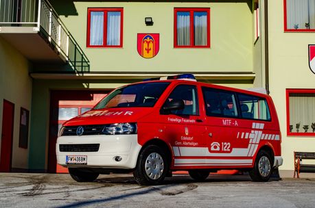Ein rotes Volkswagen-Fahrzeug, beschriftet mit 'Freiwillige Feuerwehr' und 'MTF-A', steht vor einem Gebäude mit roten Fenstern und einem gelben Emblem. Das Fahrzeug hat ein Nummernschild mit der Aufschrift 'FW 36BM'.