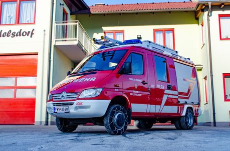 Ein roter Feuerwehrwagen mit einer Leiter auf dem Dach ist vor einem Haus mit Balkon und rot umrandeten Fenstern geparkt.