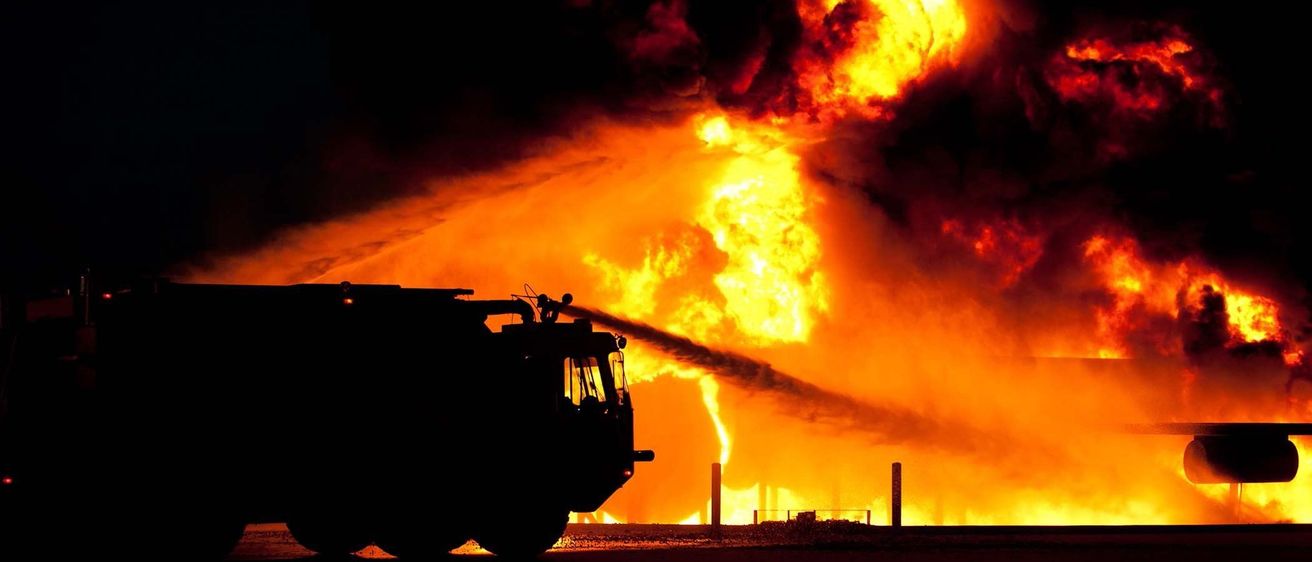 Ein Feuerwehrfahrzeug sprüht Wasser auf ein großes Feuer mit dickem Rauch. Die Szene spielt nachts.