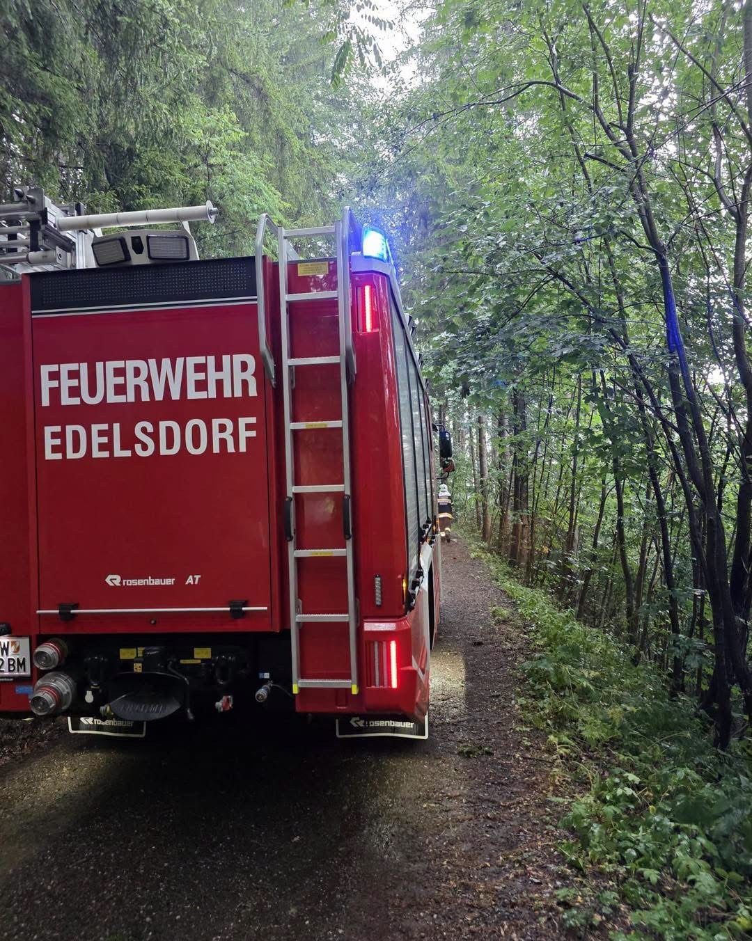 Ein roter Feuerwehrwagen mit der Aufschrift 'FEUERWEHR EDELSDORR' steht auf einem schmalen Pfad im Wald. Die Notleuchten des Fahrzeugs sind eingeschaltet.