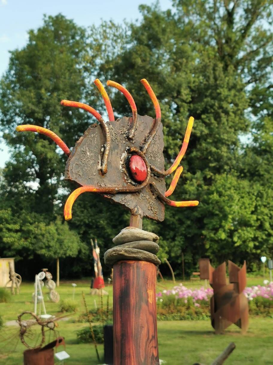 Eine abstrakte Skulptur mit einem roten Mittelpunkt steht auf einem Holzpfahl in einem Garten mit Bäumen und Blumen.