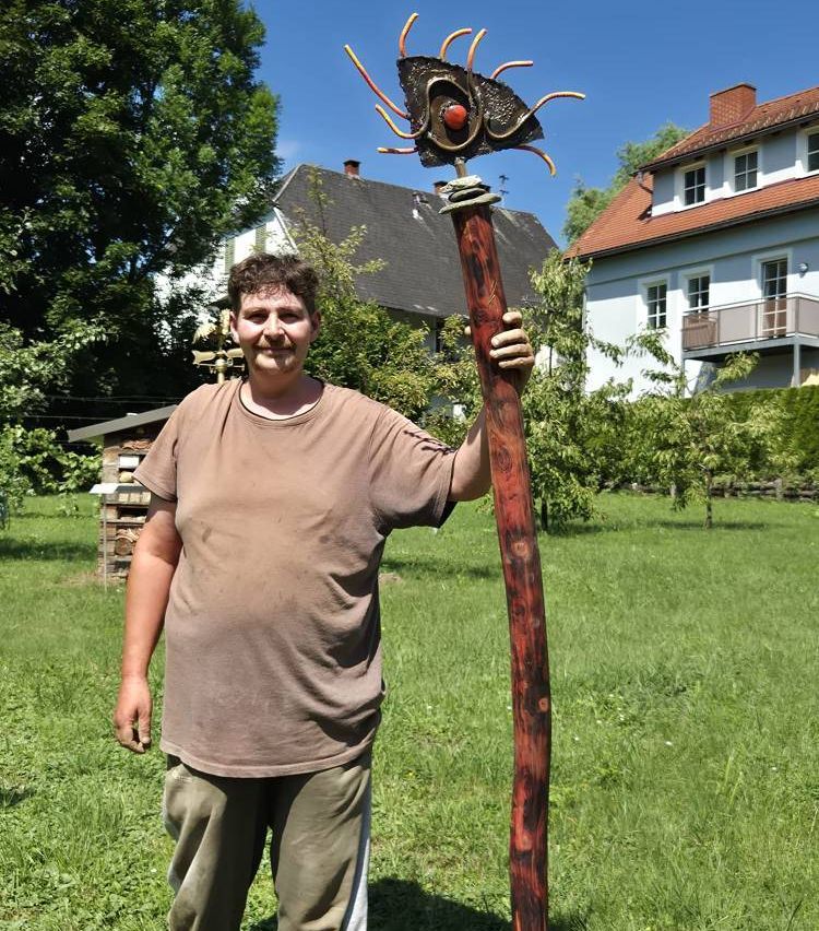 Eine Person steht in einem Garten und hält einen langen Holzstab mit einem Insektendesign an der Spitze. Dahinter befindet sich ein Haus mit Balkon und Schornstein.