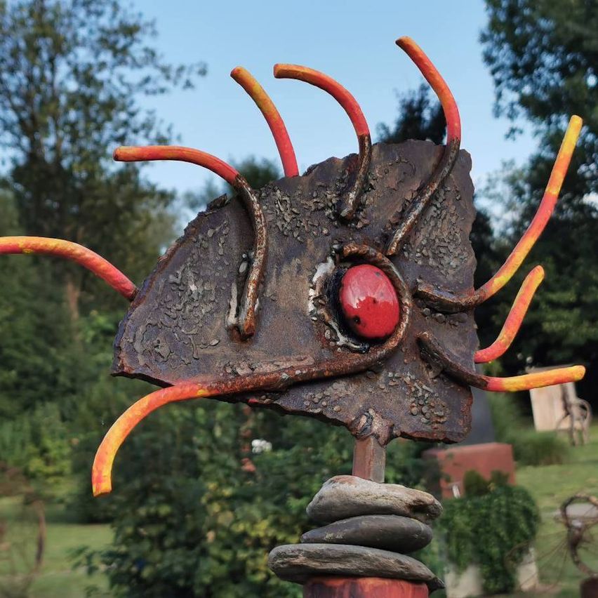 Eine Metallskulptur mit einem roten Auge und orangefarbenen, gelben und roten Stacheln steht auf Steinen. Sie steht in einem Garten mit grünen Bäumen und einem klaren Himmel.