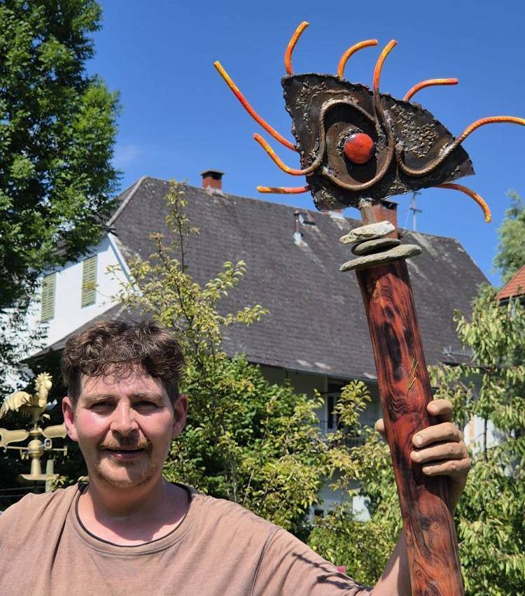 Ein Mann steht in einem Garten und hält eine einzigartige Skulptur. Sie hat ein rotes Auge und orangefarbene Ranken, die auf einem Holzstab sitzen. Hinter ihm ist ein Haus mit einem grauen Dach und weißen Fensterläden zu sehen.